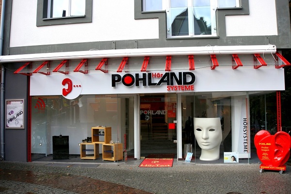 Schaufenster eines Hörakustik-Fachgeschäfts mit großem Maskenmodell und Werbeschildern.